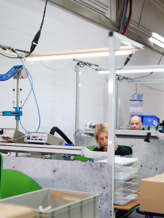 Frau mit blonden Haaren sitzt an Arbeitsplatz in Fabrikhalle mit technischen Geräten und Kartons.