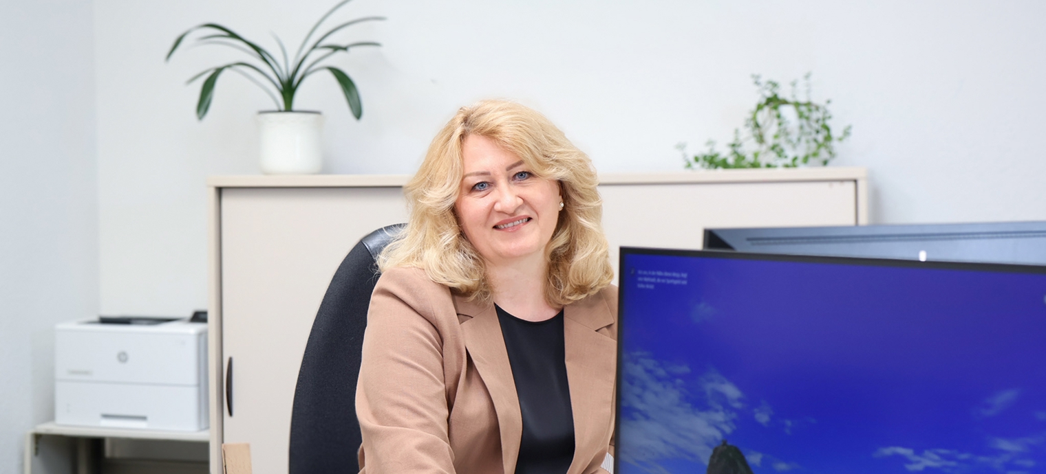 Frau mit blonden Haaren in beigem Blazer sitzt an einem Schreibtisch mit großem Monitor und Pflanzen im Hintergrund