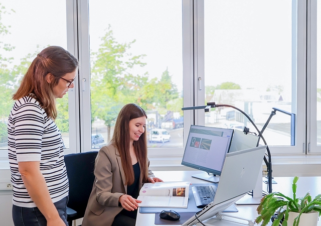 Zwei Frauen an einem Schreibtisch mit Computer und aufgeschlagenem Buch vor großen Fenstern mit Blick ins Grüne
