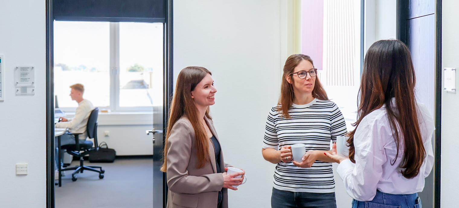 Drei Frauen stehen in einem Raum, zwei halten weiße Tassen, eine trägt ein gestreiftes Shirt, eine andere ein helles Hemd, die dritte einen Blazer.
