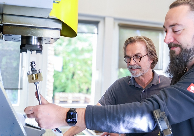 Zwei Männer bedienen gemeinsam eine gelbe CNC-Maschine in einem hellen Raum mit Fenstern