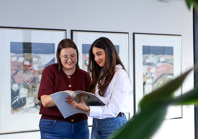 Zwei Frauen stehen nebeneinander und betrachten gemeinsam ein aufgeschlagenes Buch in einem Raum mit gerahmten Bildern an der Wand