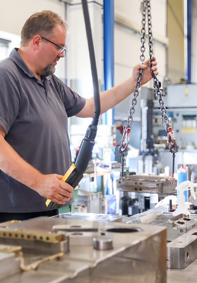 Mann in grauem Poloshirt bedient Hebekette in industrieller Fertigungshalle mit Metallteilen auf Arbeitstisch