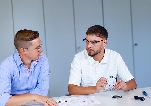 Zwei Männer sitzen an einem Tisch mit technischen Zeichnungen und Bauteilen und führen eine Diskussion