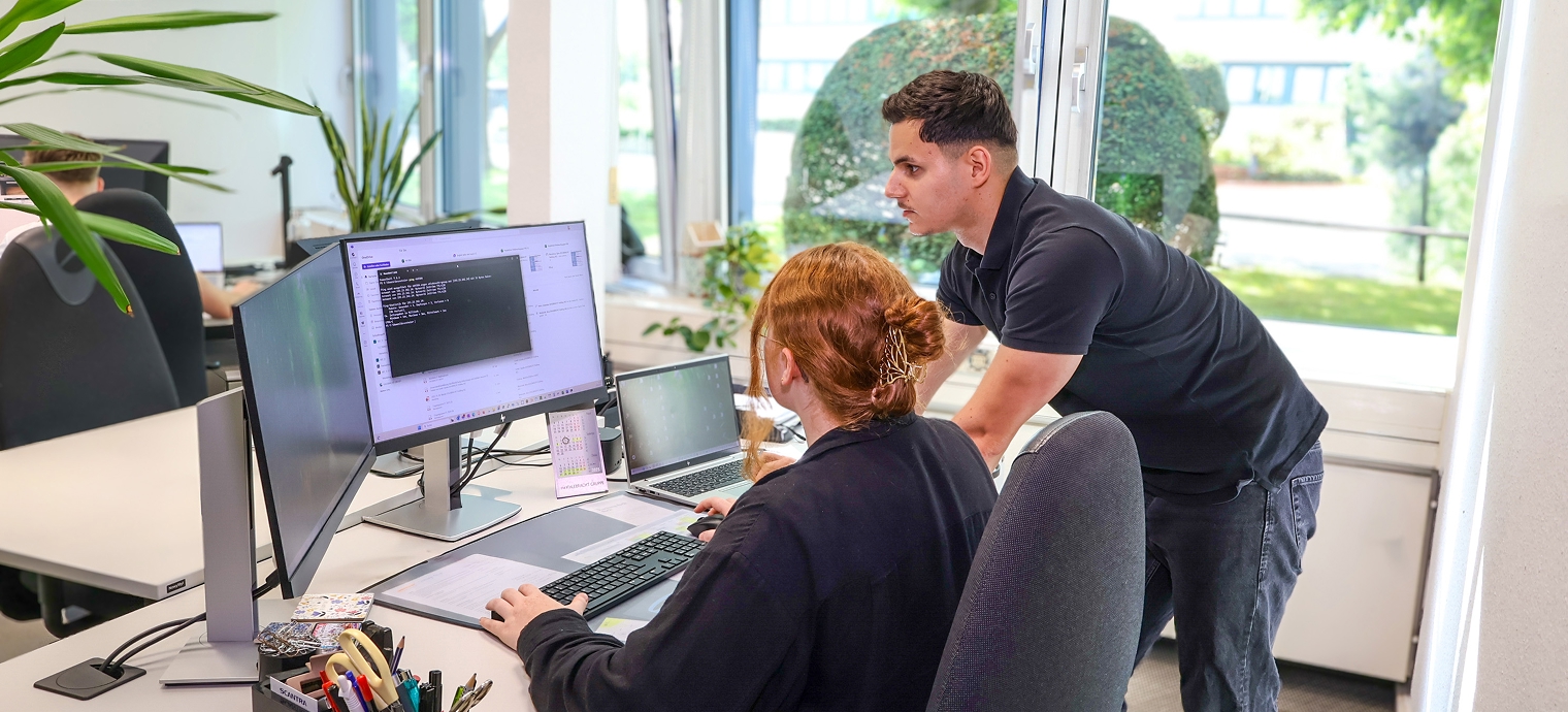 Zwei Personen an einem Schreibtisch mit zwei Monitoren, eine sitzt und tippt auf der Tastatur, die andere steht und beugt sich vor