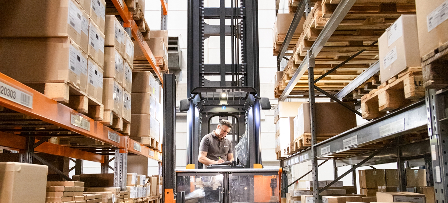 Person bedient Gabelstapler in Lager mit hohen Regalen voller Kartons und Paletten