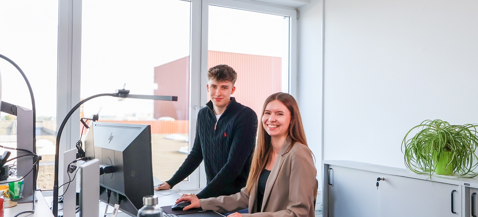 Zwei Personen an einem Schreibtisch mit Computer in einem hellen Büro mit großen Fenstern und einer Pflanze auf einem Sideboard