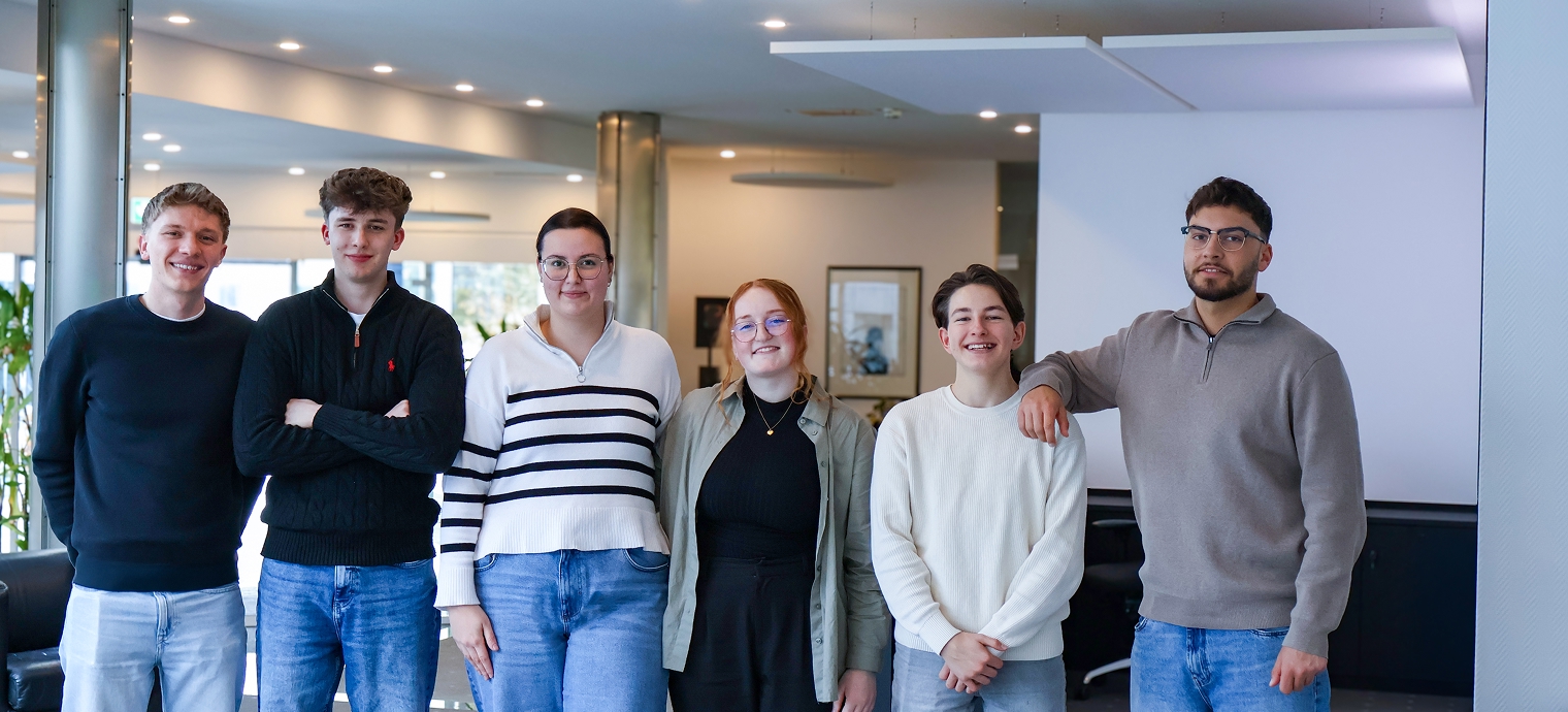 Sechs junge Erwachsene stehen nebeneinander in einem modernen Büro, einige mit verschränkten Armen, andere mit Händen in den Taschen oder auf der Schulter des Nachbarn.