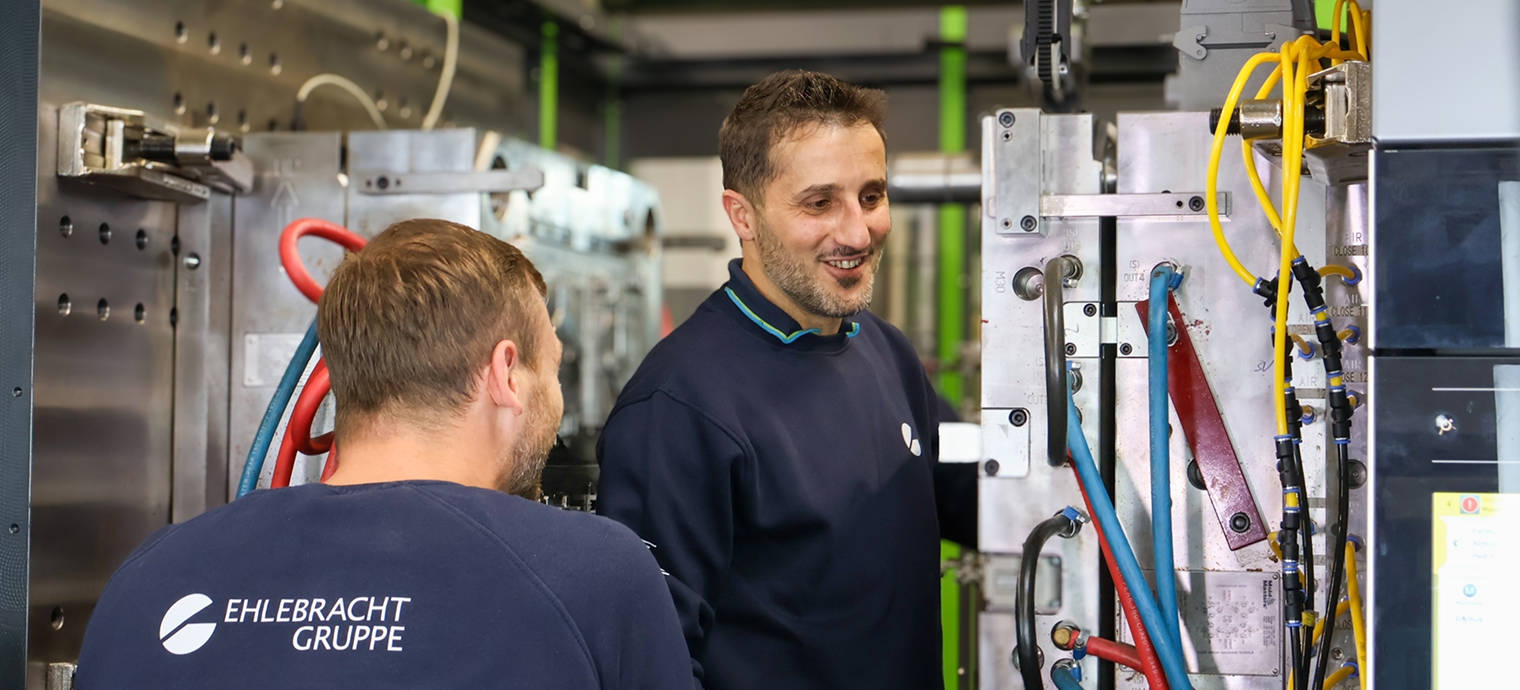 Zwei Männer in Arbeitskleidung stehen vor einer großen Maschine in einer Werkstatt oder Fabrikumgebung