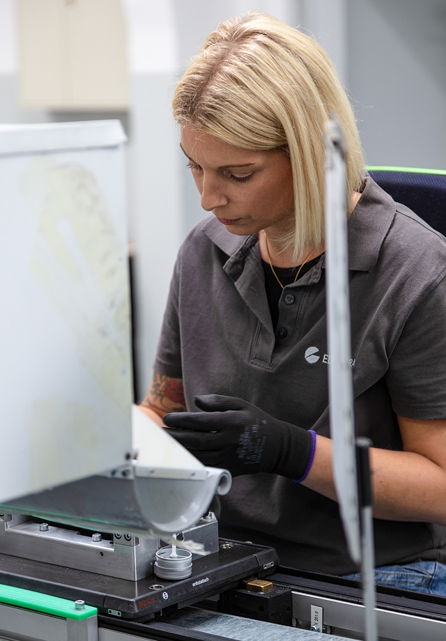 Frau mit blonden Haaren trägt schwarze Handschuhe und arbeitet an einem technischen Gerät in einer Werkstatt