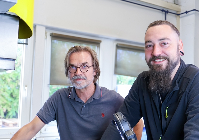 Zwei Männer in Arbeitskleidung stehen in einem hellen Raum mit großen Fenstern und Maschinen im Hintergrund