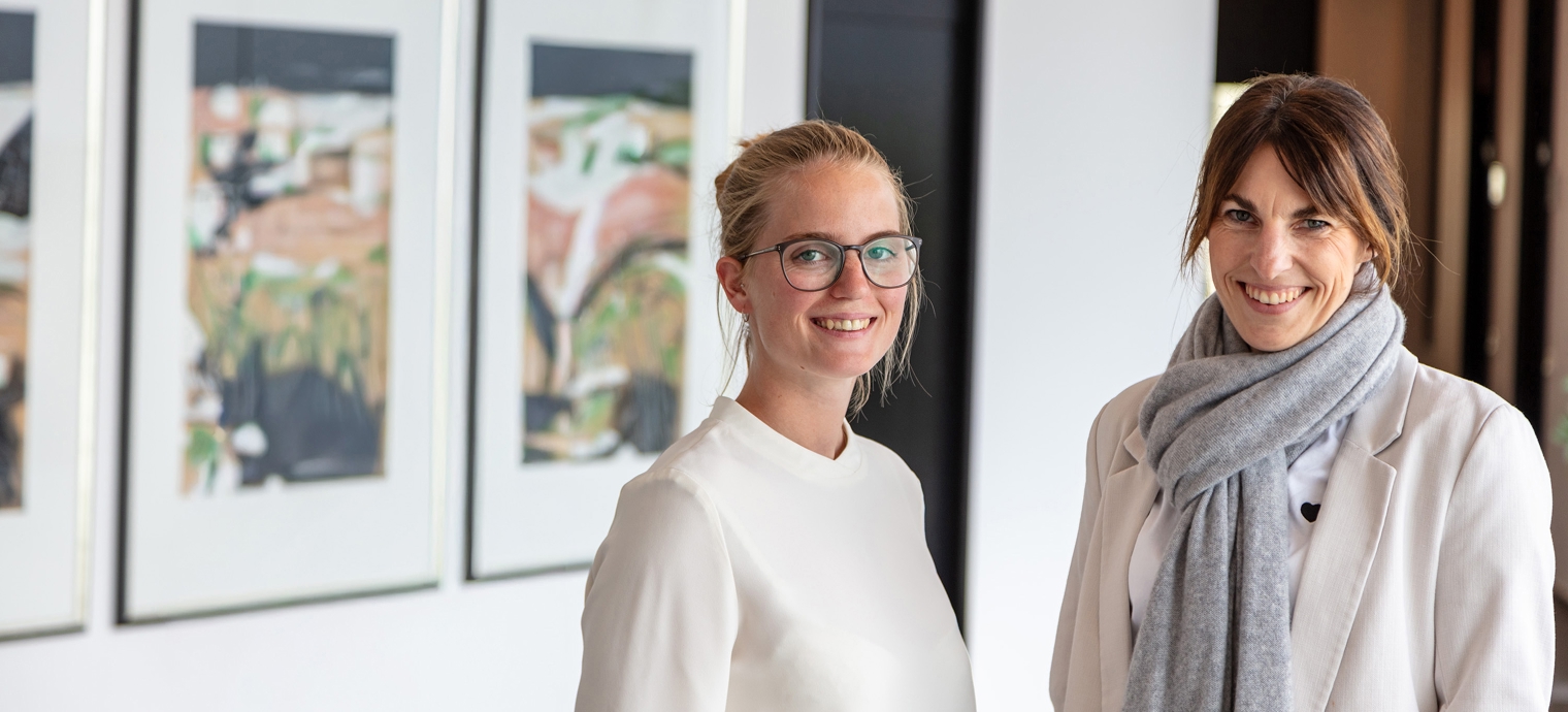 Zwei Frauen in heller Kleidung stehen vor einer weißen Wand mit drei gerahmten Bildern