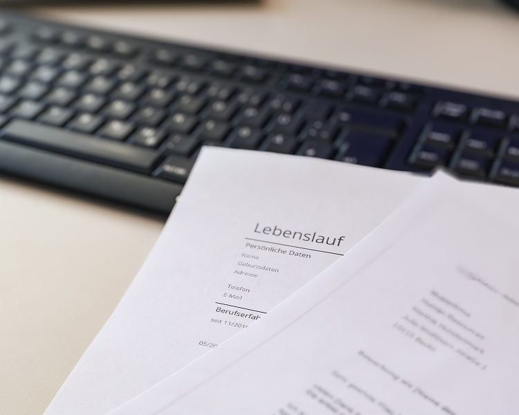 Zwei Bewerbungsunterlagen mit der Überschrift "Lebenslauf" liegen vor einer schwarzen Computertastatur auf einem Schreibtisch