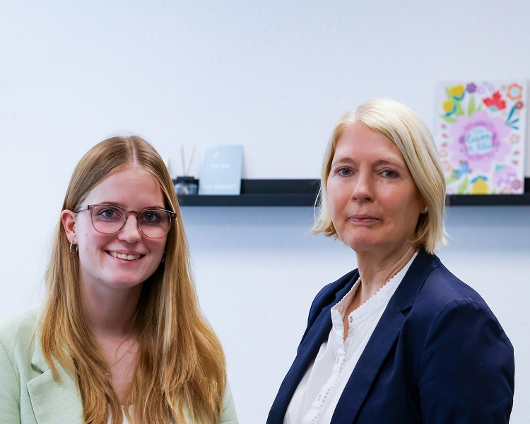 Zwei Frauen stehen nebeneinander vor einer weißen Wand mit einem schwarzen Wandregal und Dekorationen