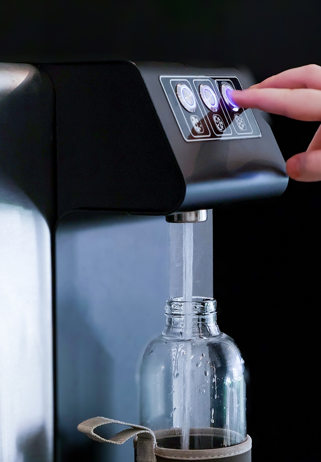 Wasserspender mit Glasflasche, aus der Wasser fließt, während eine Hand eine beleuchtete Taste am Bedienfeld drückt