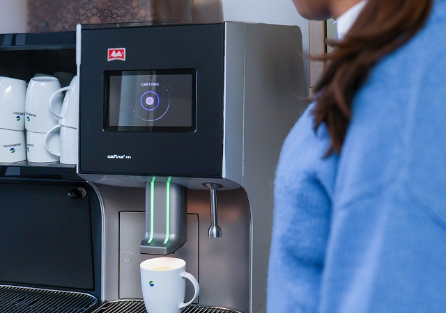 Kaffeemaschine mit digitalem Display und Tasse darunter, davor steht eine Person im blauen Pullover