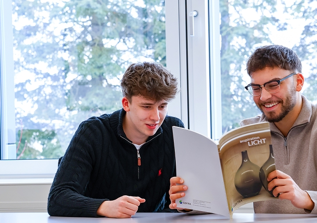 Zwei Männer sitzen an einem Tisch vor einem Fenster, einer hält einen Katalog mit dem Titel 'Inspiration LICHT'