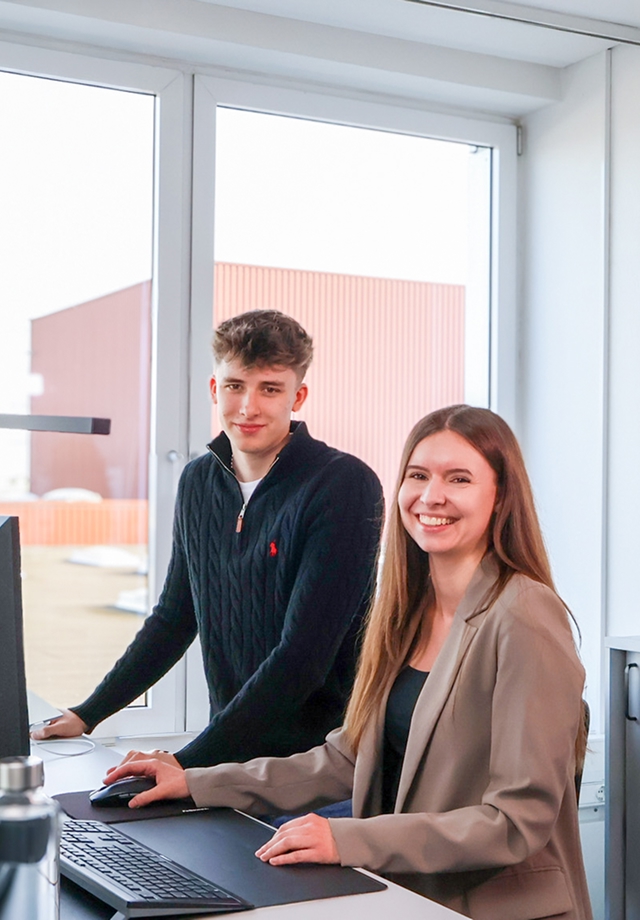 Zwei Personen an einem Schreibtisch mit Computer vor groĂźen Fenstern in einem hellen BĂĽro