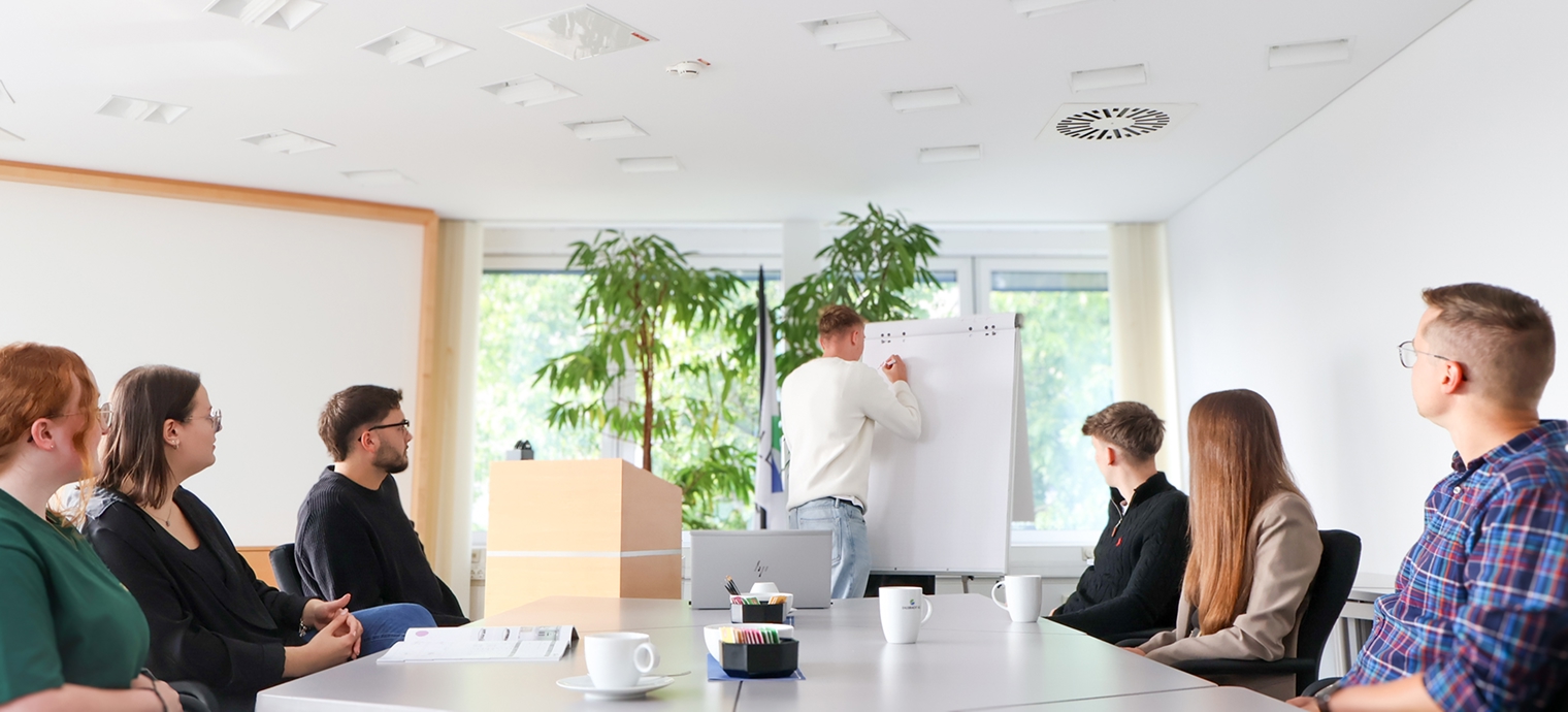 Person steht vor Flipchart und schreibt, mehrere Personen sitzen an Konferenztisch und schauen zu