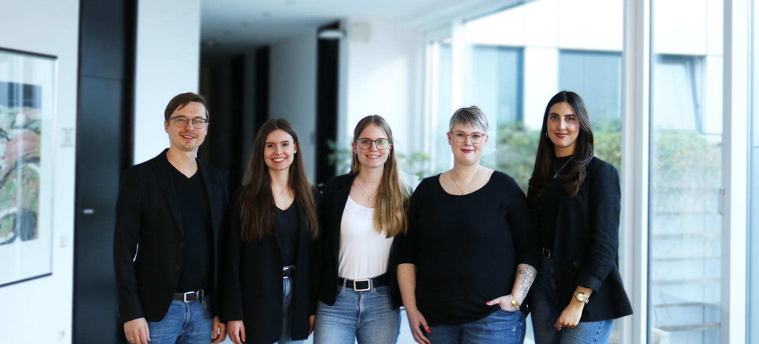 Fünf Personen stehen nebeneinander in einem modernen Bürogebäude, alle tragen legere Kleidung mit Jeans und Oberteilen, zwei Personen tragen Blazer.