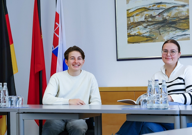 Zwei Personen sitzen an einem Tisch mit mehreren Wasserflaschen, im Hintergrund stehen drei Fahnen und an der Wand hängt ein abstraktes Gemälde